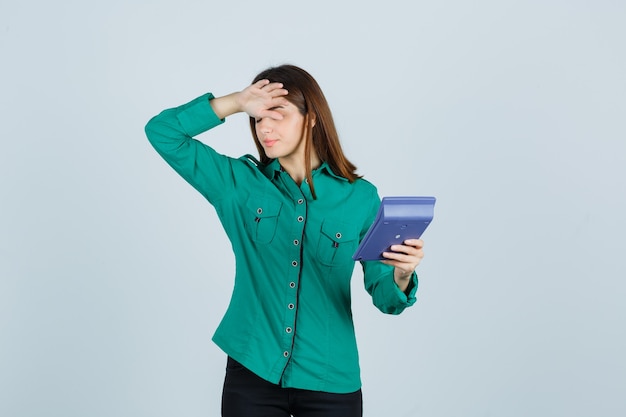 Jeune femme tenant la calculatrice tout en gardant la main sur le front en chemise verte et à la frustration. vue de face.
