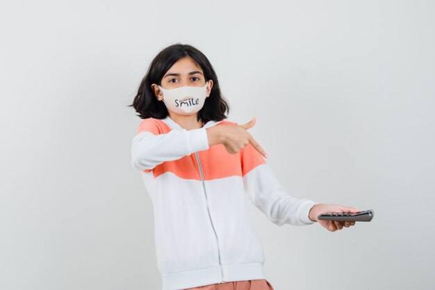 Jeune femme tenant la calculatrice tout en montrant le pouce vers le haut dans un sweat à capuche, un masque et un air heureux.