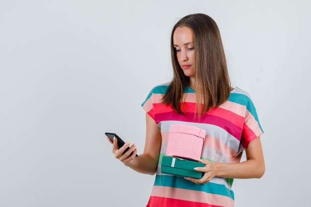Jeune femme tenant des coffrets cadeaux et regardant le téléphone en vue de face de t-shirt.