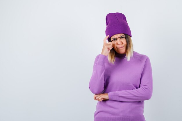 Jeune femme tenant le doigt sur la tête en pull violet, bonnet et semblant sérieuse, vue de face.