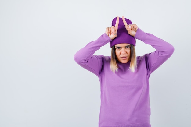 Jeune femme tenant les doigts au-dessus de la tête comme des cornes de taureau en pull violet, bonnet et l'air amusé, vue de face.