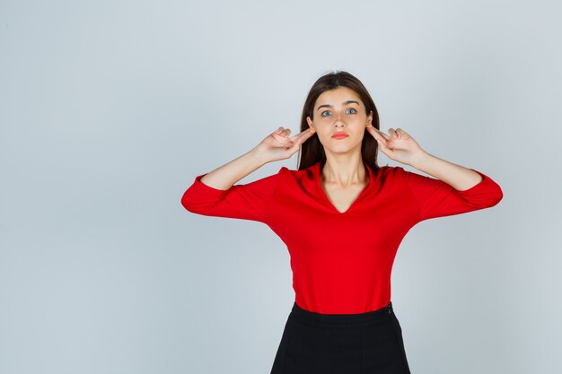 Jeune femme tenant les doigts derrière les oreilles en chemisier rouge, jupe et à la sérieuse