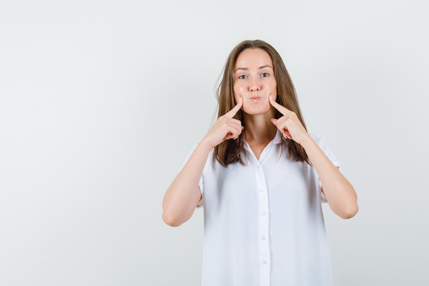 Photo gratuite jeune femme tenant les doigts ses joues en chemisier blanc et à étrange