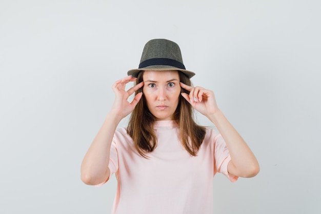 Photo gratuite jeune femme tenant les doigts sur les tempes en t-shirt rose, chapeau et à la sérieuse. vue de face.