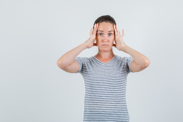 Jeune femme tenant les doigts sur la tête en t-shirt et à la confusion, vue de face.