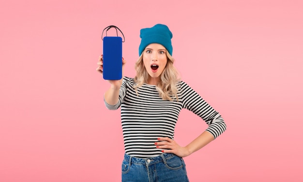 Jeune femme tenant haut-parleur sans fil écouter de la musique portant une chemise rayée et un chapeau bleu souriant posant sur rose