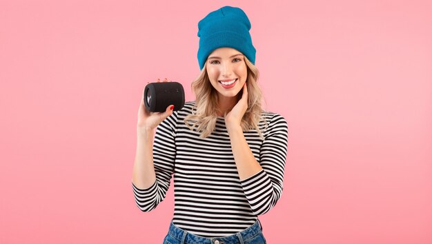 Jeune femme tenant haut-parleur sans fil écouter de la musique portant une chemise rayée et un chapeau bleu souriant posant sur rose