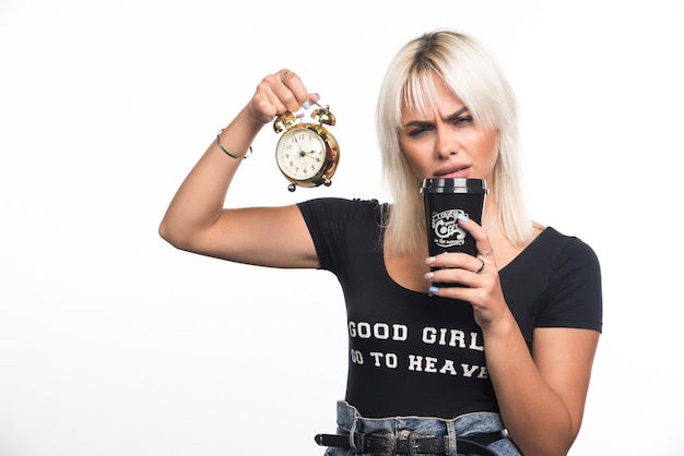 Jeune femme tenant une horloge et en prenant une gorgée de tasse de café sur un mur blanc.