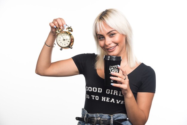 Jeune femme tenant une horloge et une tasse de café sur un mur blanc.