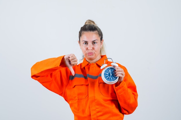 Photo gratuite jeune femme tenant une horloge tout en montrant le pouce vers le bas en uniforme de travailleur et à la mécontentement. vue de face.
