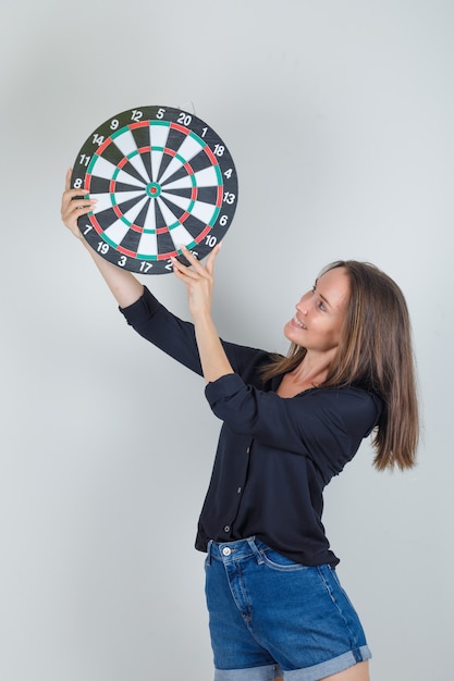 Photo gratuite jeune femme tenant un jeu de fléchettes en chemise noire, short en jean et à la joyeuse