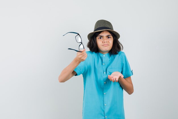 Jeune femme tenant des lunettes en chemise bleue, chapeau et à la recherche d'impuissance