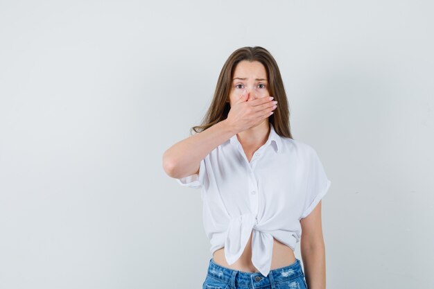 Jeune femme tenant la main sur la bouche en chemisier blanc et à la peur. vue de face.