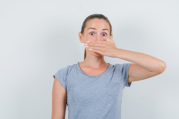 Photo gratuite jeune femme tenant la main sur la bouche en t-shirt gris et à la peur. vue de face.