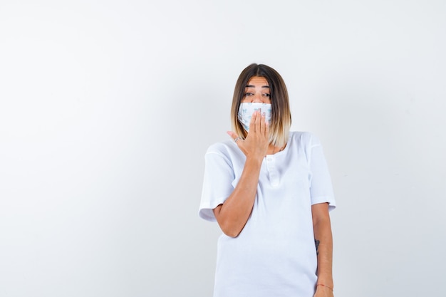 Jeune femme tenant la main sur la bouche en t-shirt, masque et à la joyeuse, vue de face.