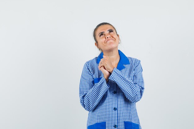 Jeune femme tenant la main dans la pose de prière, rêvant de quelque chose en chemise de pyjama vichy bleu et à la jolie. vue de face.