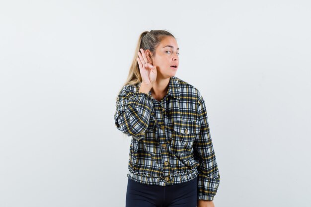 Jeune femme tenant la main derrière l'oreille en chemise, short et à la curieuse, vue de face.