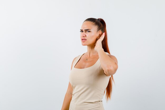 Jeune femme tenant la main derrière l'oreille en débardeur beige et à la recherche concentrée. vue de face.