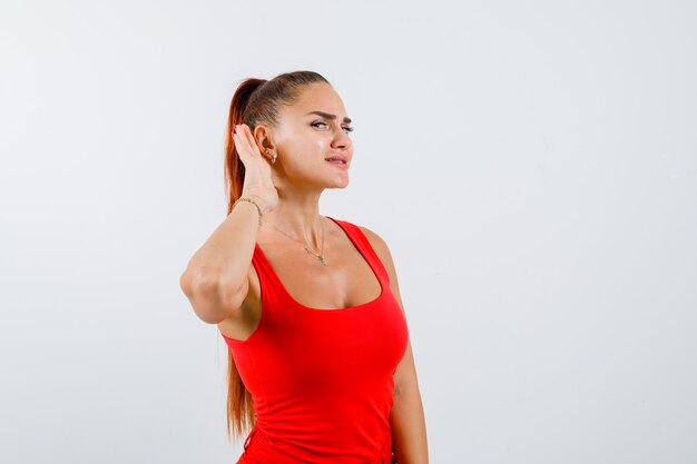 Jeune femme tenant la main derrière l'oreille en débardeur rouge, pantalon et à la curieuse, vue de face.