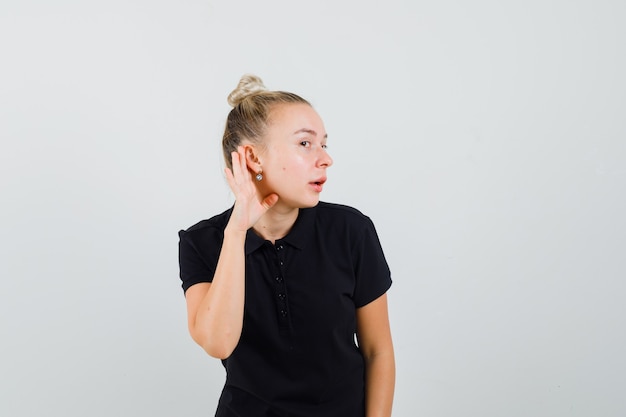 Jeune femme tenant la main derrière l'oreille en t-shirt noir et à la curieuse. vue de face.