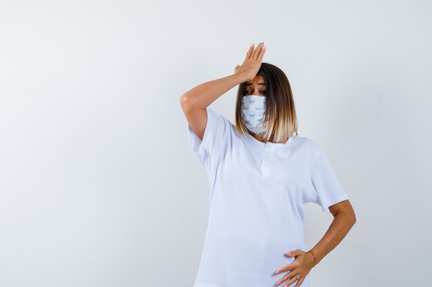 Jeune femme tenant la main sur le front en t-shirt, masque et à la vue oublieuse, de face.