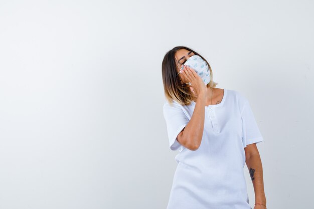 Jeune femme tenant la main sur la joue tout en regardant ailleurs en t-shirt, masque et à la vue réfléchie, de face.