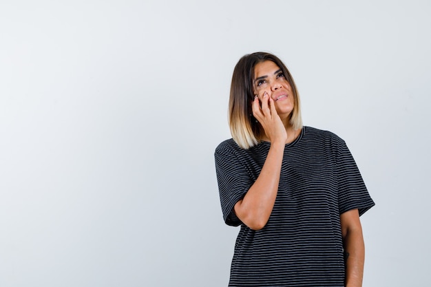 Jeune femme tenant la main sur la joue tout en regardant en robe de polo et à la jolie. vue de face.