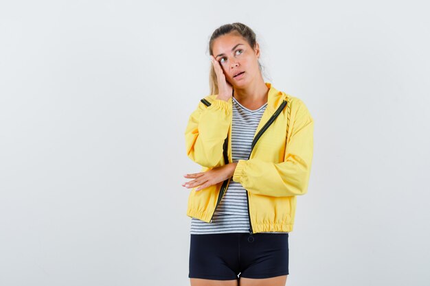 Jeune femme tenant la main sur la joue en veste, t-shirt et à la rêveuse, vue de face.