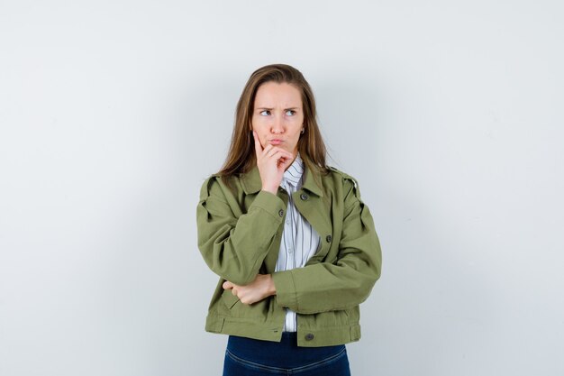 Jeune femme tenant la main sur le menton en chemise, veste et hésitante, vue de face.