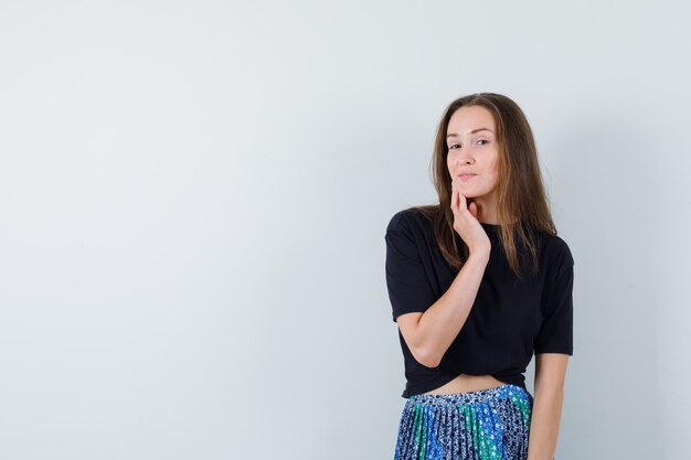 Jeune femme tenant la main sur le menton et posant à la caméra en t-shirt noir et jupe bleue et à la charmante