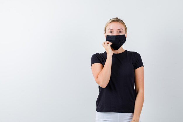 Jeune femme tenant la main sur le menton en t-shirt, pantalon, masque médical et l'air perplexe