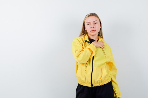 Jeune femme tenant la main sur la poitrine en veste jaune, pantalon et à la vue de face, confiant.
