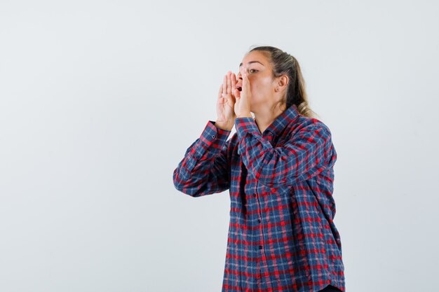 Jeune femme tenant la main près de la bouche comme appelant quelqu'un en chemise à carreaux et à la jolie