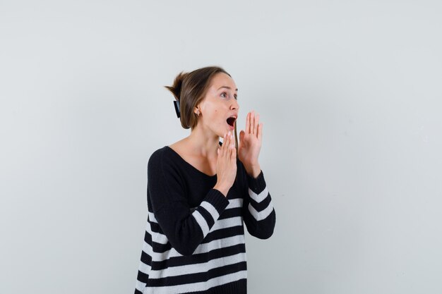 Jeune femme tenant la main près de la bouche ouverte en chemise et à la recherche d'excitation