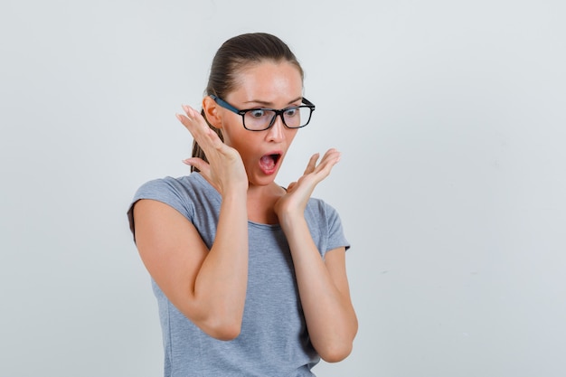 Jeune femme tenant la main près de la bouche en t-shirt gris, lunettes et à la peur. vue de face.