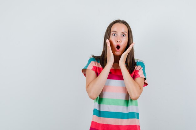 Jeune femme tenant la main près de la bouche en t-shirt et à la vue choquée, de face.