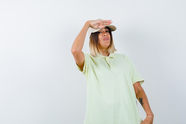 Jeune femme tenant la main près du capuchon pour voir clairement en t-shirt, casquette et à la confiance. vue de face.