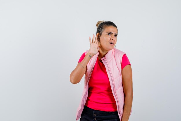 Jeune femme tenant la main près de l'oreille pour entendre quelque chose en t-shirt rose et veste et à la concentration