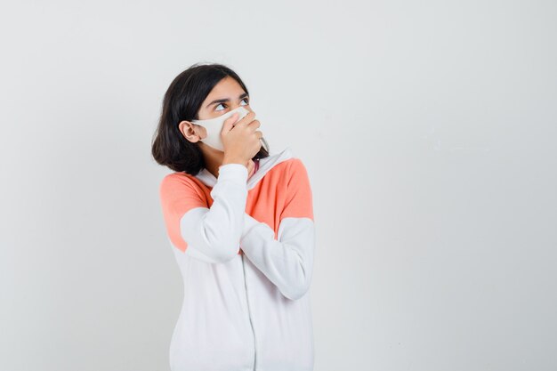 Jeune femme tenant la main sur sa bouche en sweat à capuche, masque facial et à la peur.