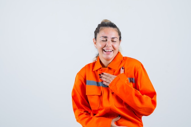 Jeune femme tenant la main sur sa poitrine en riant en uniforme de travailleur et à la joyeuse vue de face.