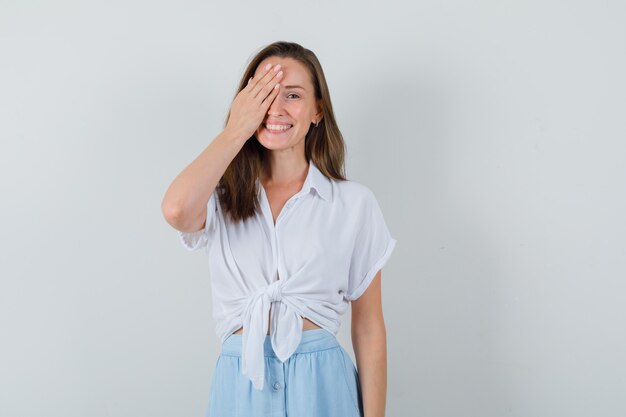 Jeune femme tenant la main sur son œil tout en souriant en chemisier, jupe et à la ludique