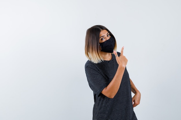 Jeune femme tenant une main sur la taille, pointant vers le haut en robe noire, masque noir et à la grave, vue de face.