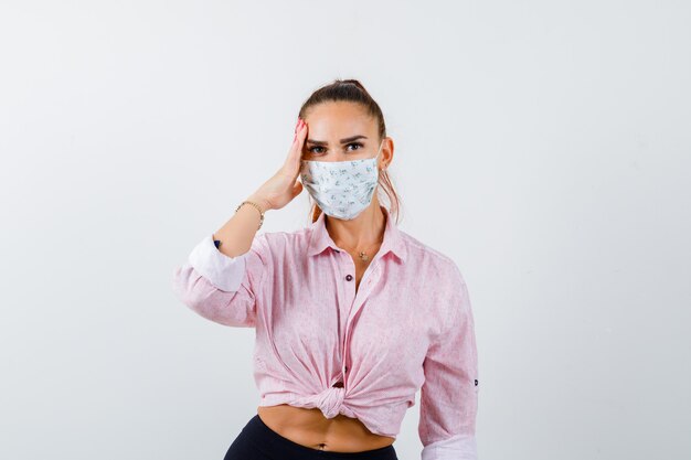 Jeune femme tenant la main sur la tête en chemise, pantalon, masque médical et à la vue réfléchie, de face.