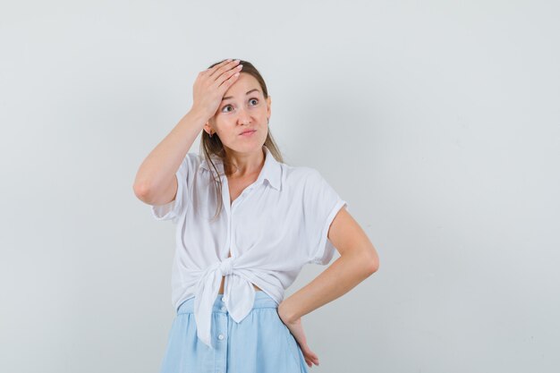 Jeune femme tenant la main sur la tête en chemisier et jupe et à la recherche de désolé