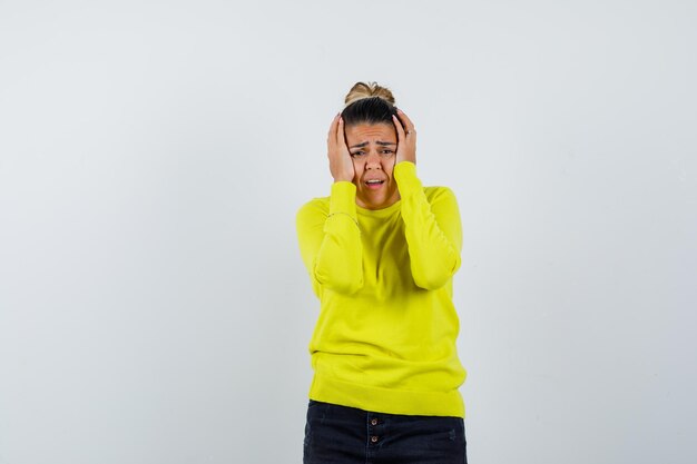 Jeune femme tenant la main sur la tête en pull jaune et pantalon noir et à la harcelé