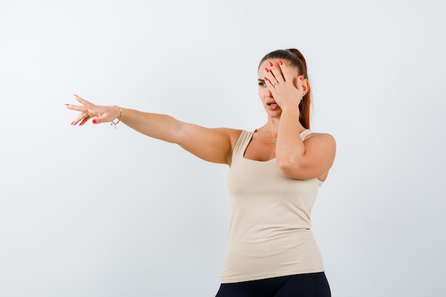 Jeune femme tenant la main sur les yeux tout en étirant le bras en débardeur beige et à la grave, vue de face.