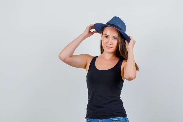 Jeune femme tenant les mains sur le chapeau et souriant en maillot, vue de face de short.