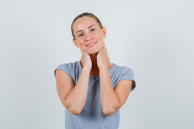 Jeune femme tenant les mains sur le cou en t-shirt gris et à la joyeuse vue de face.