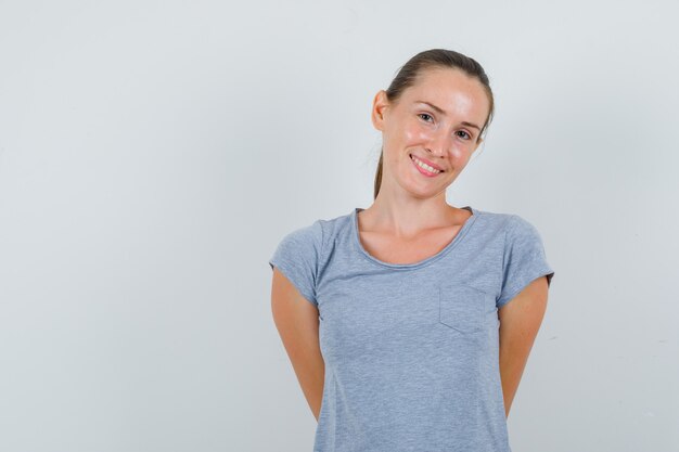 Jeune femme tenant les mains sur le dos en t-shirt gris et à la joyeuse. vue de face.