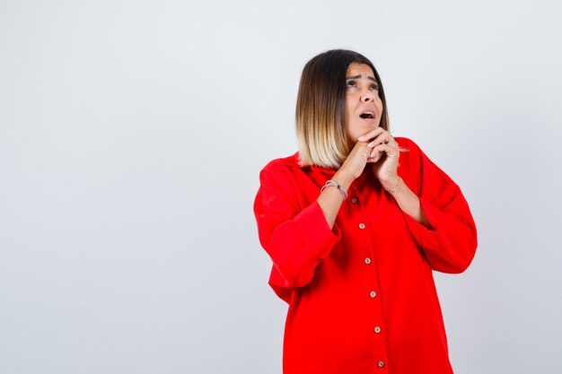 Jeune femme tenant les mains jointes sous le menton en chemise rouge surdimensionnée et à la perplexité, vue de face.